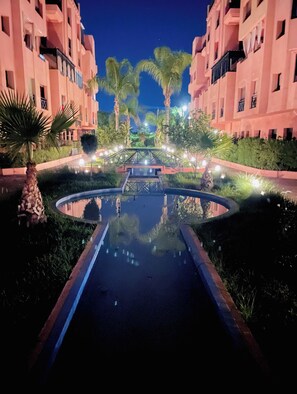 Ein Hotel nachts, mit einer zentralen Wasserspiele, Palmen und beleuchteten Wegen.