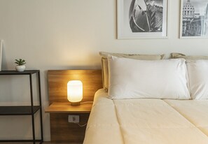 A neatly made bed with white linens, a bedside lamp, and a small potted plant on a nightstand.