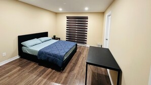 Ein Schlafzimmer mit Bett, kleinem Tisch, Fenster mit Jalousien und Tür.