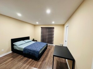 A bedroom with a bed, a small table, a window with blinds, and a door.