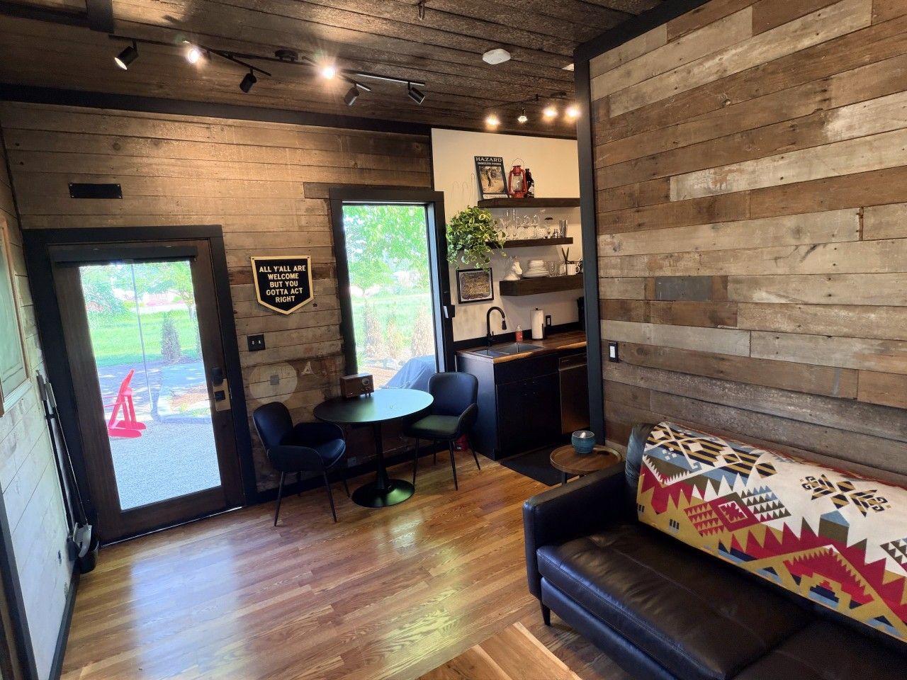 A modern interior with a wooden wall, a sofa, a dining area with chairs, and a kitchen area with a sink and shelves.