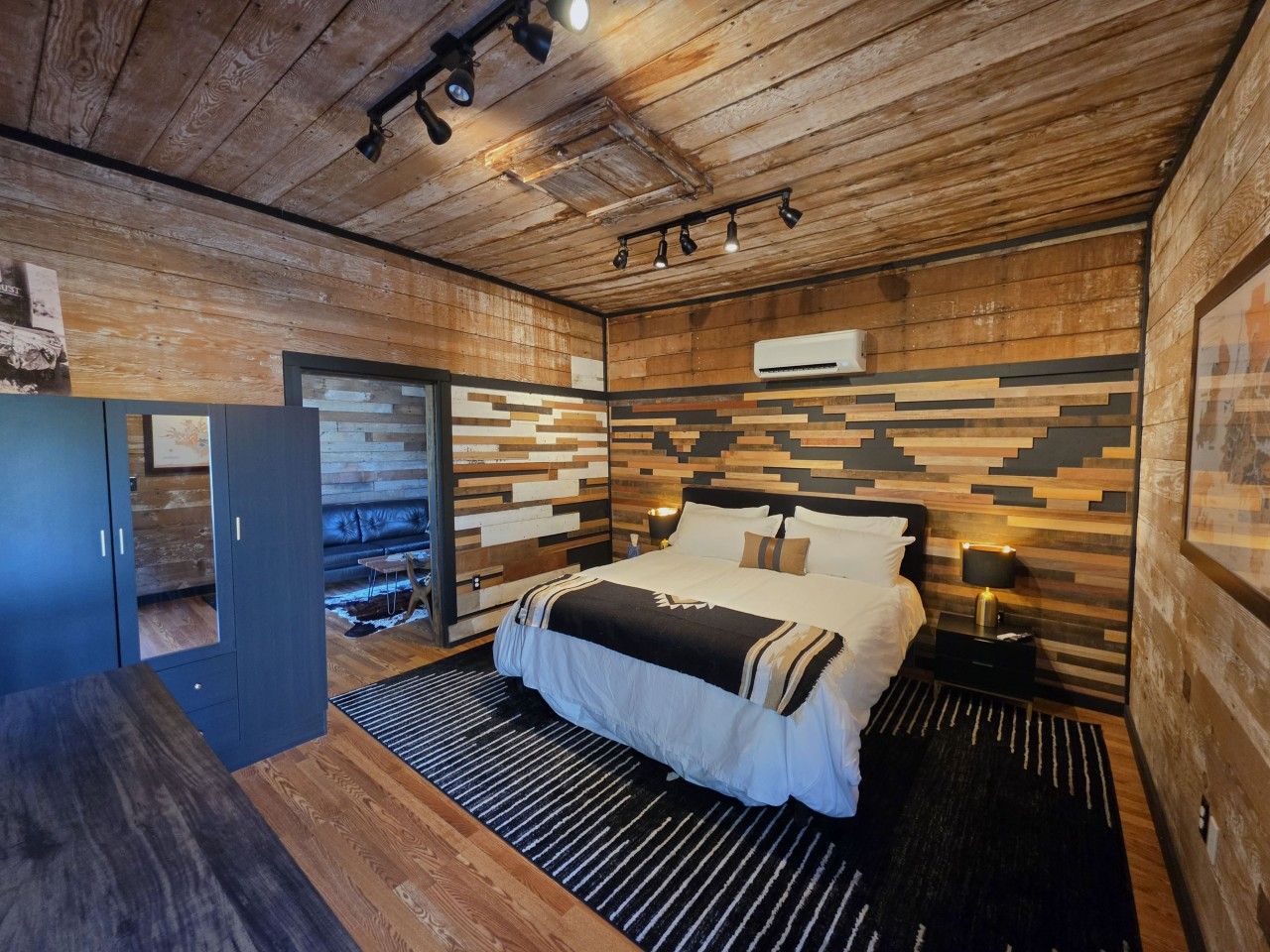 A modern bedroom with a wooden ceiling, a bed with a striped bedspread, a nightstand with a lamp, and a wall-mounted air conditioner.
