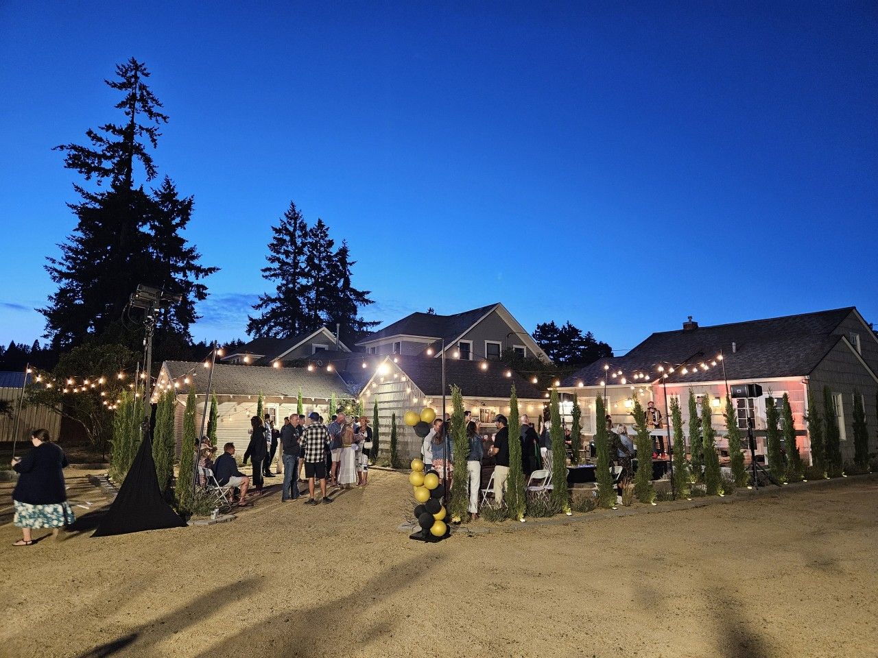 An outdoor event with people gathered around a decorated area, tents, and string lights.
