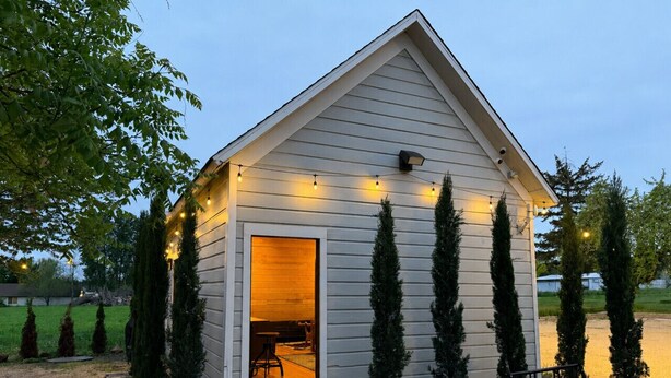 A small, single-story building with a sloped roof, a door leading inside, and string lights along the exterior.