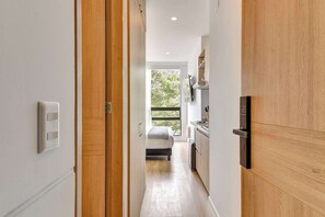 Een smalle gang die uitkomt in een slaapkamer met een bed, een raam en een kitchenette.