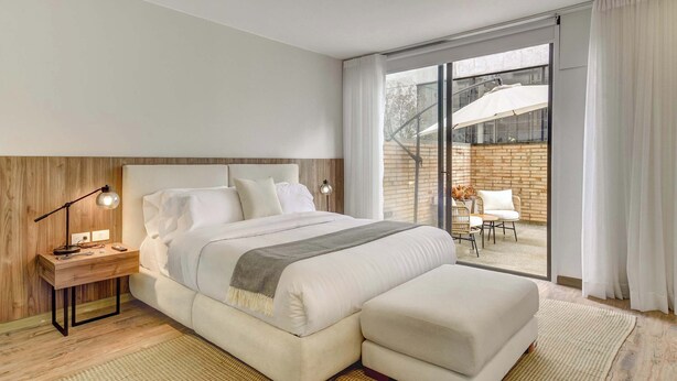 Een moderne hotelkamer met een groot bed, een nachtkastje met lamp, een schuifdeur naar een balkon en uitzicht op de omgeving.