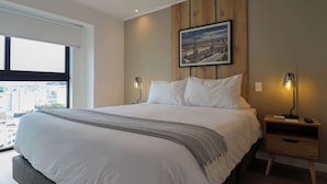 A bedroom with a large bed, a cityscape painting on the wall, a nightstand with a lamp, and a view of buildings outside the window. - w* | Cityview 2BR w/ Balcony in Miraflores (Miraflores)