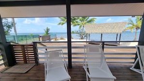 Una terraza techada con dos sillones blancos, piso de madera, un paraguas de paja y vista al océano y palmeras.