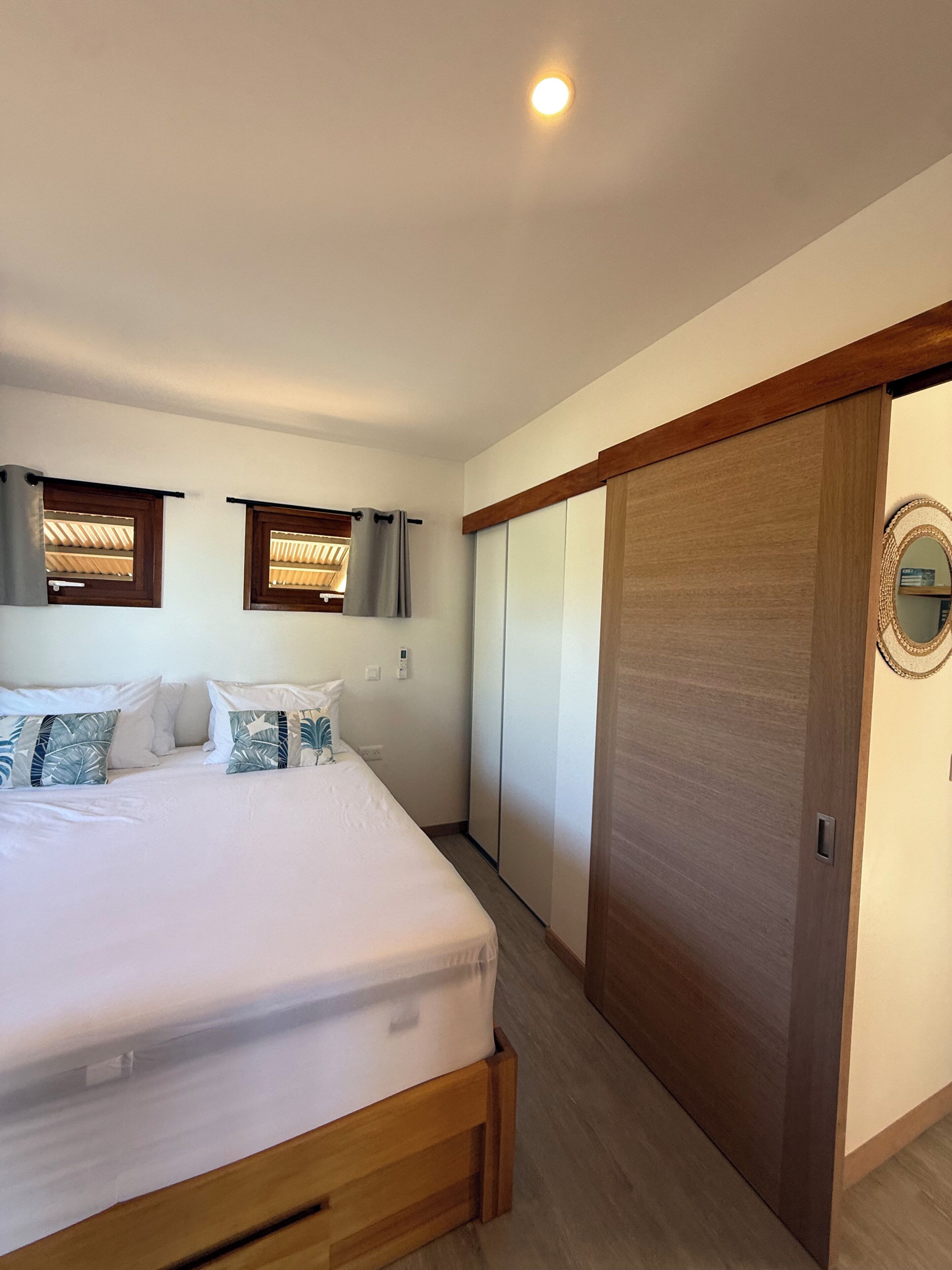 A bedroom with a bed, two windows with curtains, a wooden door, and a mirror on the wall.