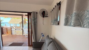 A modern room with a grey sofa, a ceiling fan, and a view of a beach. - Le TeRava (Raiatea)