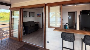 A modern interior with a wooden deck, a bar area with stools, a sofa, and a kitchen visible through an open door. - Le TeRava (Raiatea)