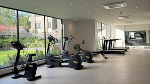 A gym with stationary bikes, treadmills, and a large window offering a view of greenery and buildings. - Delali Residence Luxury 1BR Near Airport, Pool, Gym & private Terrace (Accra)
