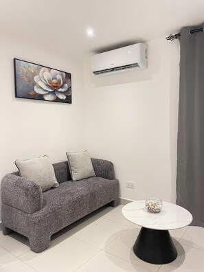 A modern living room with a grey sofa, a round black table, and a wall-mounted air conditioner. - Delali Residence Luxury 1BR Near Airport, Pool, Gym & private Terrace (Accra)
