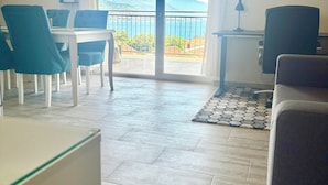 Quarto com mesa de jantar, cadeiras, sofá, escrivaninha com abajur e vista para uma varanda com guarda-corpo e montanhas ao fundo.