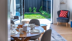 Een eethoek met een tafel gedekt voor twee personen, uitzicht op de achtertuin en een grijze fauteuil met een rood kussen.