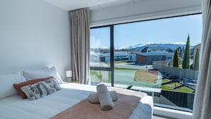 Hotelkamer met een groot raam, een bed, nachtkastje en uitzicht op een gebouw en bergen.