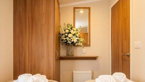 A hotel room with two beds, a wooden wardrobe, a mirror, a radiator, and a bouquet of flowers on a shelf. - 3BR Premium Caravan | Near Edinburgh |Parking|WiFi (Port Seton)