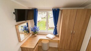 A compact room with a wooden vanity, a mirror, a television, a window with curtains, and a wardrobe. - 3BR Premium Caravan | Near Edinburgh |Parking|WiFi (Port Seton)