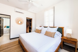 A hotel room with a bed, a nightstand, a wall-mounted air conditioner, a ceiling fan, and a window.
