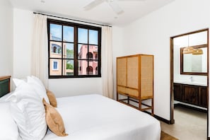 Una habitación de hotel con cama, ventana con cortinas, armario de ratán, espejo y ventilador de techo.