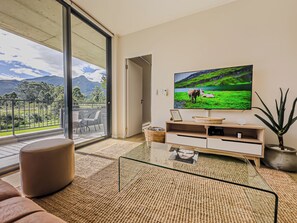 Uma sala de estar moderna com uma televisão grande, uma mesa de centro de vidro e vista para uma paisagem verde através de uma porta de correr.