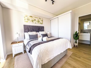 Un dormitorio moderno con cama, mesitas de noche, un tocador, un ventanal grande y un baño con lavamanos y espejo.