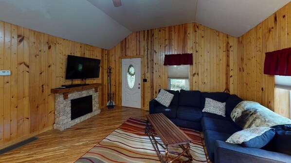 A cozy cabin interior with wooden walls, a stone fireplace, a dark blue sofa, and a television on the wall.