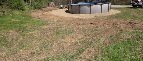Een rond zwembad met een kiezelpad eromheen, bomen op de achtergrond en een geparkeerd voertuig in de buurt.