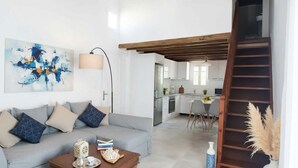 Un salon moderne avec un canapé, une table basse et des œuvres d’art. On aperçoit une cuisine à l’arrière-plan, avec des meubles blancs et un coin repas.