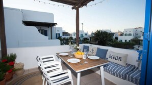 Une terrasse avec une table dressée pour deux personnes, des chaises blanches, un banc avec un coussin et une vue sur des bâtiments blancs.