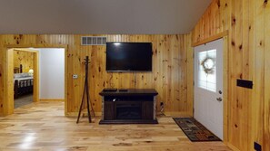 Ein Zimmer mit Holzwänden, einem Fernseher über einem Kamin, einer Tür mit Glasfenster und einem Gang zu einem weiteren Raum.