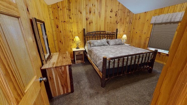 A wooden-paneled room with a bed, bedside lamps, a small wooden dresser, and a window with blinds. - Scotts Creek Cabins - Hilltop (Logan)