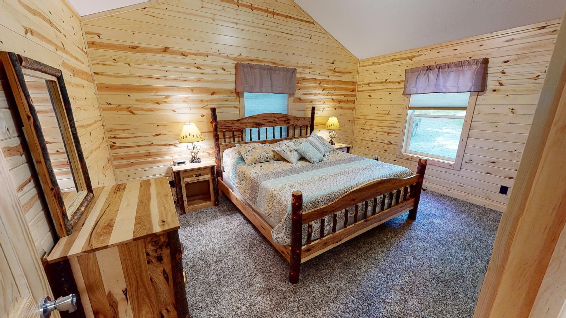 A wooden cabin bedroom with a bed, two bedside tables, a mirror, and two windows with curtains.