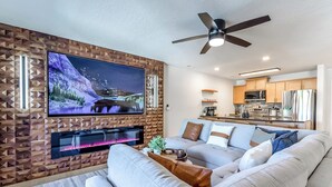 A modern living room with a flat-screen TV, a ceiling fan, and a kitchen area with wooden cabinets and stainless steel appliances. - Sunset Lakefront Retreat with Luxe Upgrades (Osage Beach)
