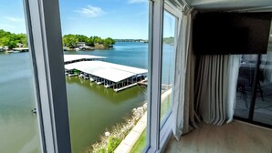 A room with a large window overlooking a calm lake, a dock with boats, and a distant bridge. - Sunset Lakefront Retreat with Luxe Upgrades (Osage Beach)
