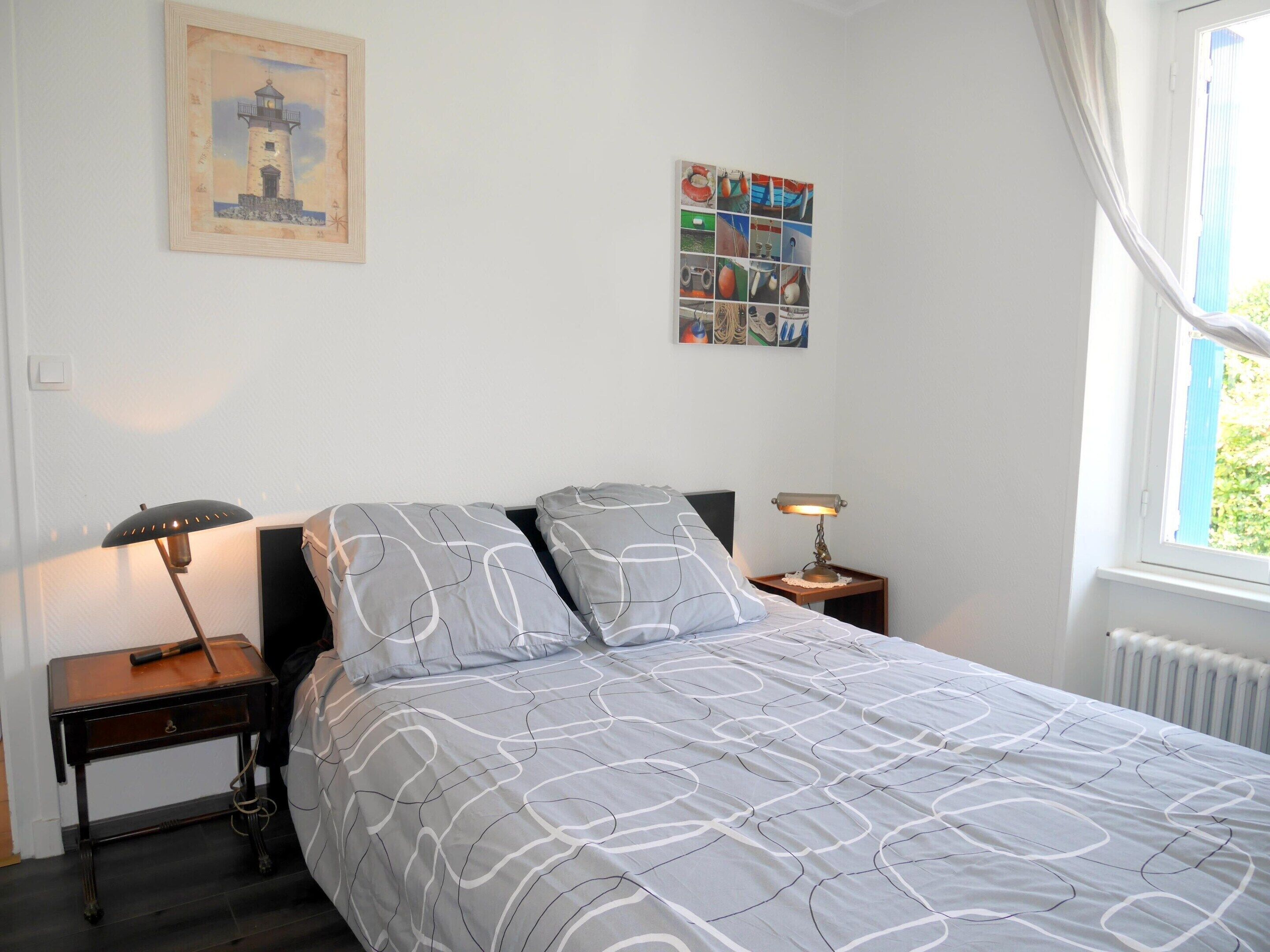 Un dormitorio con una cama, una mesita de noche, una lámpara, un cuadro enmarcado y una ventana.