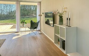 Un salón moderno con un ventanal corredero que da a un jardín, un sillón verde, una consola blanca con un jarrón y velas, y un cuadro grande en la pared.