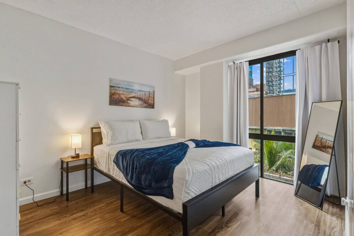A bedroom with a bed, bedside lamp, a painting, a window with curtains, and a mirror.