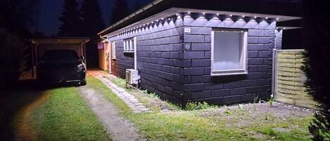 A small, dark-colored building with a window and a door, a paved driveway, and a car parked in the background.