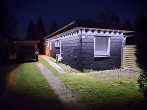A small, dark-colored building with a window and a door, a paved driveway, and a car parked in the background. - Renovated holiday home with hot tub Marielyst (Væggerløse)