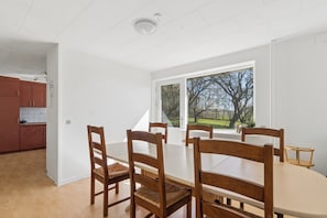 A dining area with a large table and four wooden chairs, a window with a view of trees, and a kitchen in the background.
