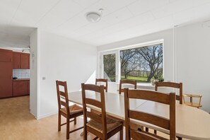 A dining area with a large table and four wooden chairs, a window with a view of trees, and a kitchen in the background. - Holiday Charm on Bornholms South Coast (Aakirkeby)