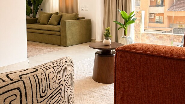 A modern living room with a sofa, a patterned ottoman, a round coffee table, and a view of a building through large windows. - 10-Mins from Cairo Airport, Garden Oasis (Cairo Governorate)