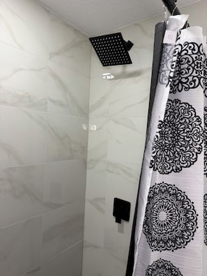 Un baño con baldosas de mármol blanco, cabezal de ducha, toallero negro y cortina de ducha estampada.