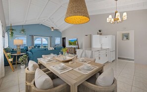 A spacious living area with a dining table set for four, a sofa, a ceiling fan, a TV, and a refrigerator.