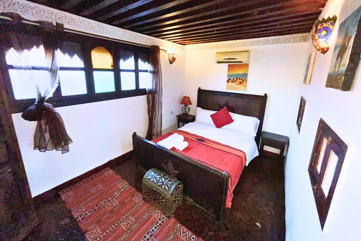A room with a bed, a nightstand, a decorative lantern, a window with curtains, and a patterned rug.
