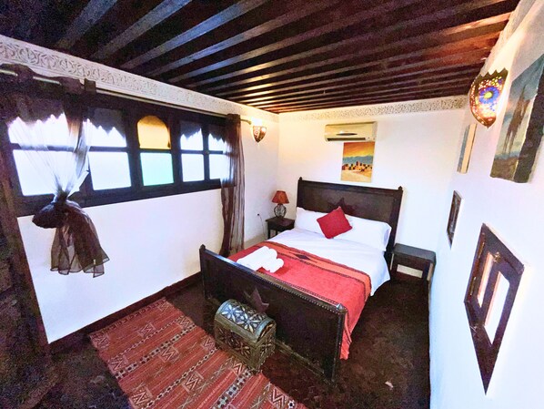 Ein Zimmer mit einem Bett, einem Nachttisch, einer dekorativen Laterne, einem Fenster mit Vorhängen und einem gemusterten Teppich.