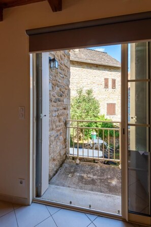 A room with a sliding glass door leading to a balcony with a stone wall, a metal railing, and a view of a building with a tiled roof. - L'OUSTAL DE LUCIE - SAINT SATURNIN (SAINT SATURNIN)