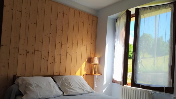 A bedroom with a wooden headboard, a bed with white bedding, a bedside lamp, and a window with sheer curtains. - Gîte Le Toura 2/4 persons (SAINT-ALBAN-SUR-LIMAGNOLE)