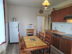 A compact kitchen with a refrigerator, microwave, wooden table, chairs, and a dishwasher. - Gîte Le Toura 2/4 persons (SAINT-ALBAN-SUR-LIMAGNOLE)